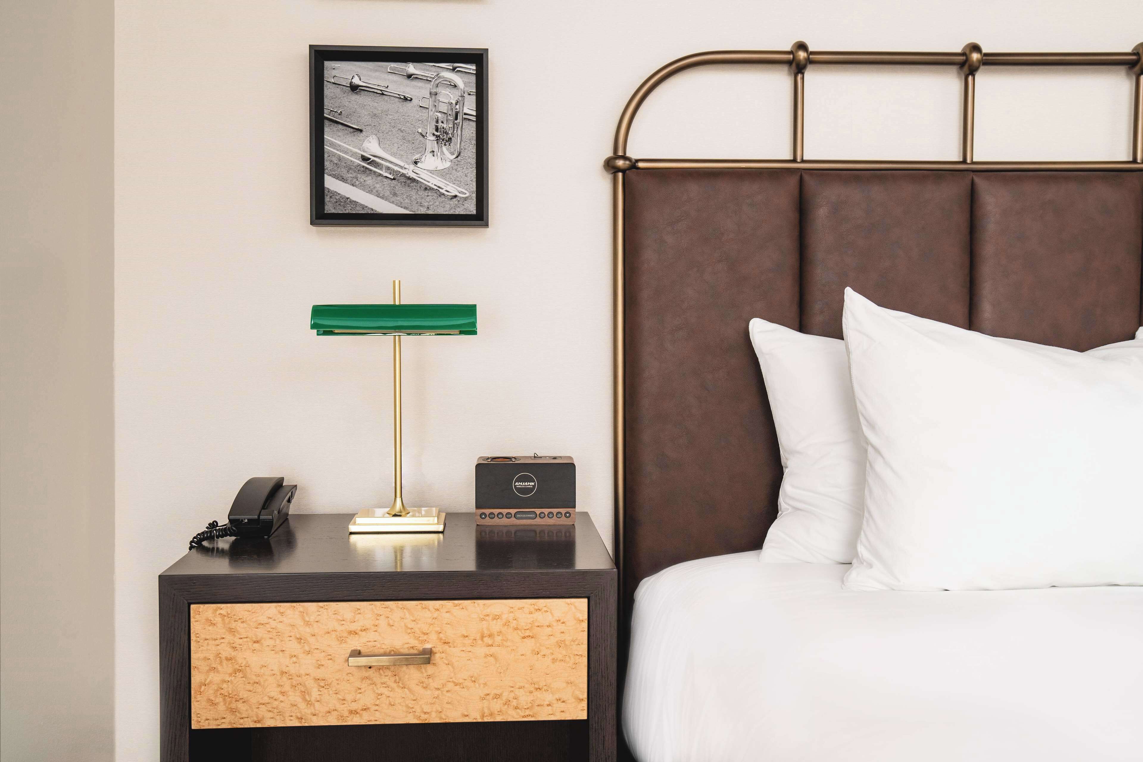 Una cama bien hecha con almohadas blancas, un cabecero oscuro, una mesita de noche con una lámpara, un teléfono y una radio pequeña.