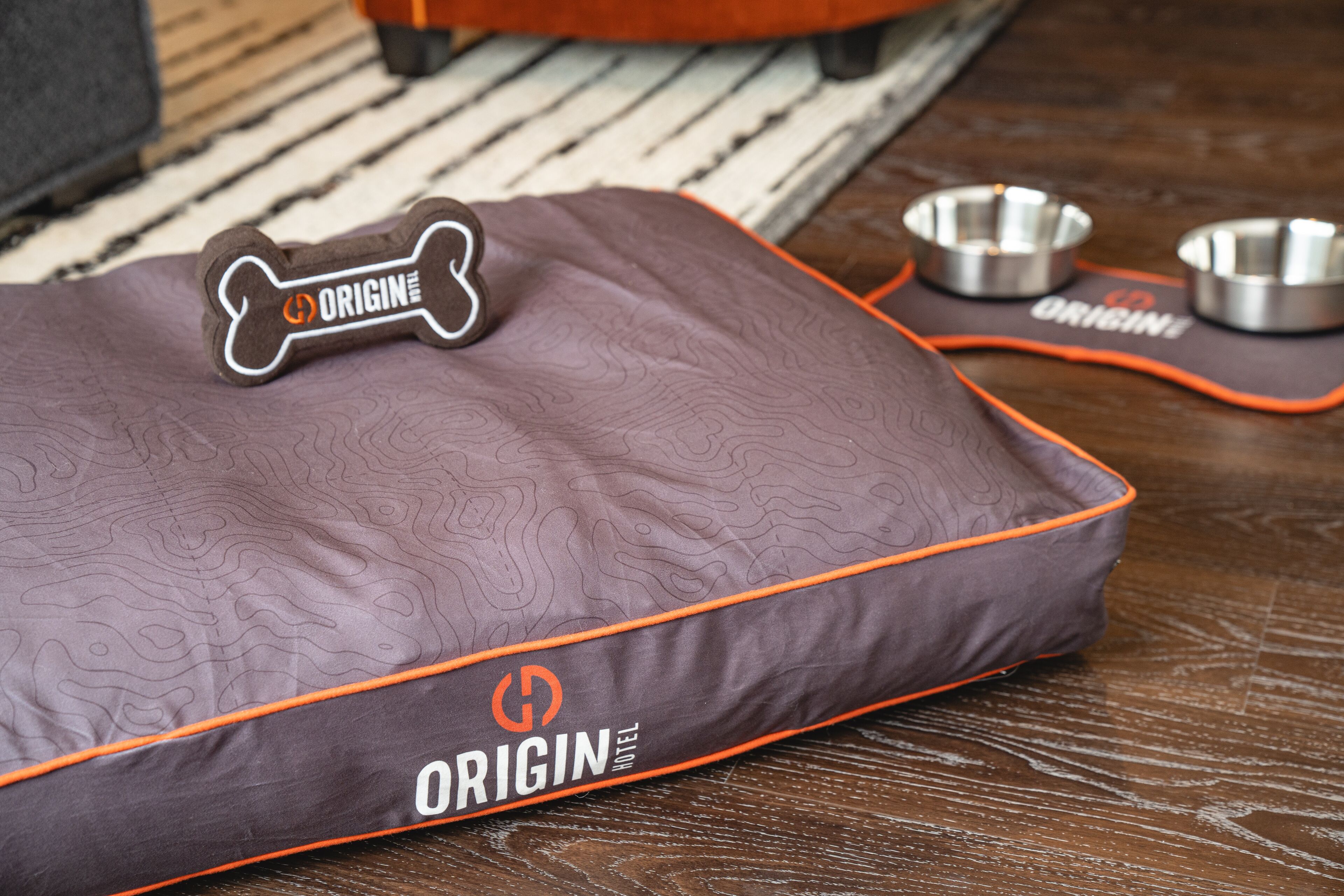 A pet bed with the 'ORIGIN HOTEL' branding, a bone-shaped tag, and two metal bowls on a wooden floor.