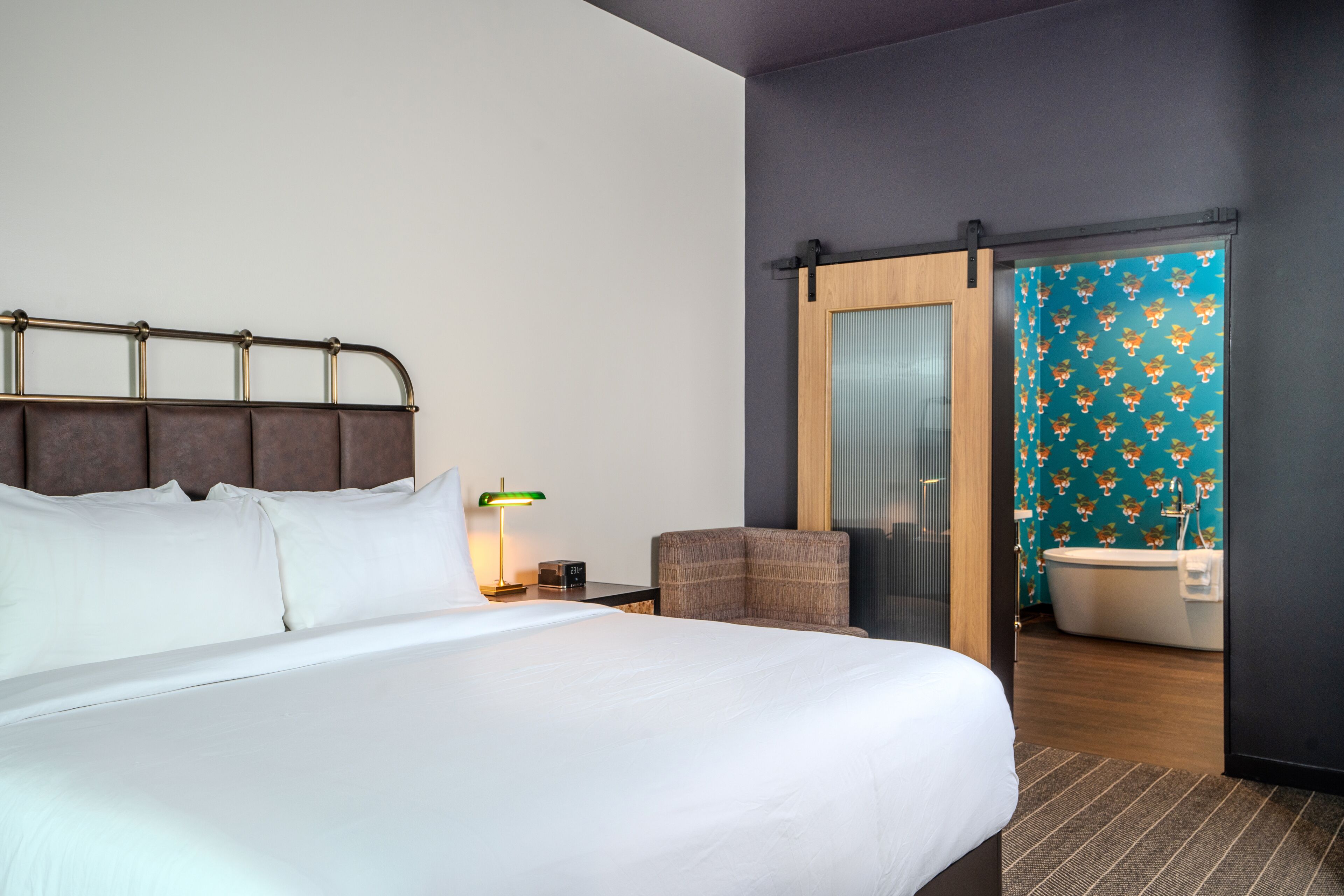 A hotel room with a large bed, a chair, a nightstand with a lamp, a wooden door, and a bathroom with a bathtub and toilet.