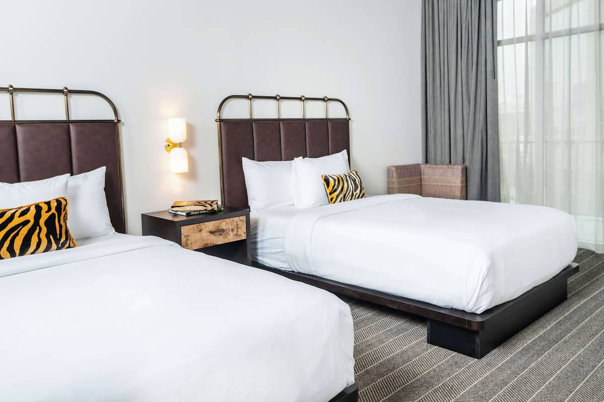 A hotel room with two beds, a nightstand, a lamp, and a view of the city through large windows.
