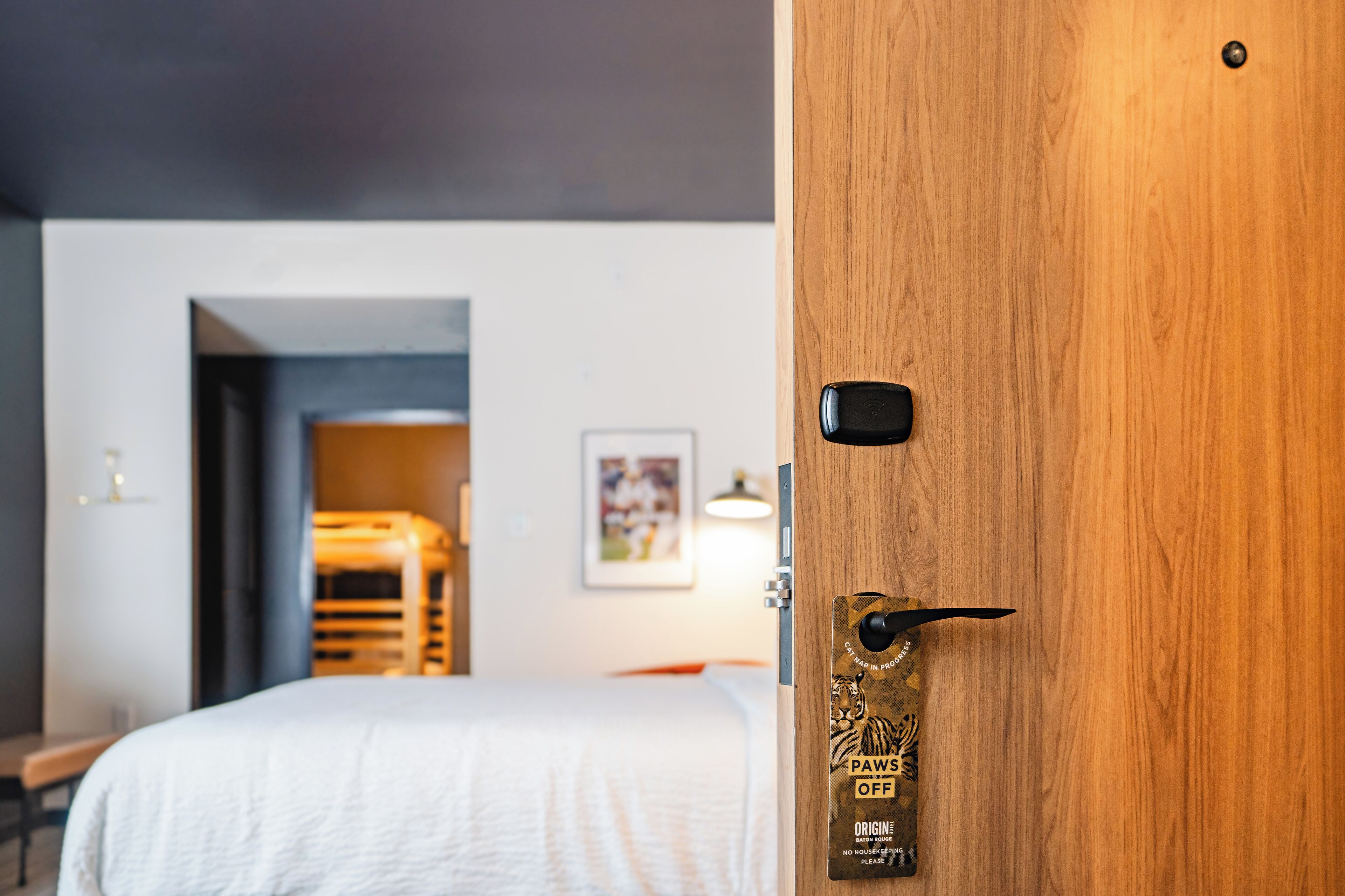 A hotel room with a wooden door, a bed with a white bedspread, a framed picture on the wall, and a small table with a lamp.