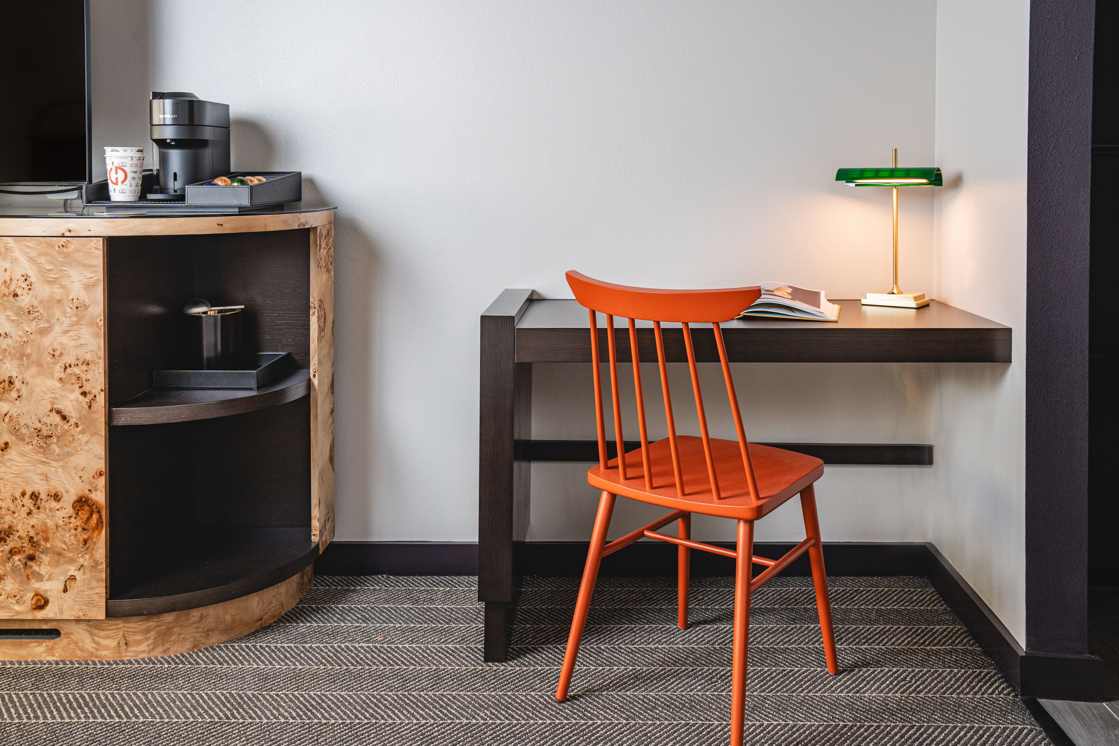 Un petit espace de travail comprenant un meuble en bois, une chaise, un bureau, une machine à café et une lampe de bureau verte.