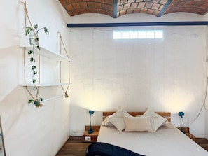 Une chambre à coucher avec un lit, deux tables de chevet, une plante suspendue et une fenêtre.