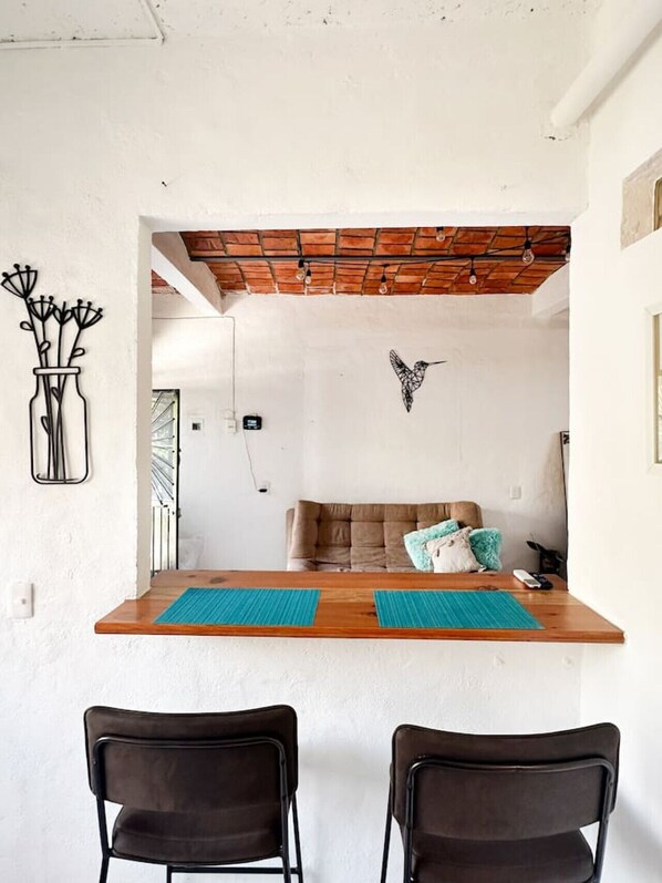 Une pièce comprenant une table en bois, deux chaises, un canapé avec des coussins, un luminaire suspendu et un élément décoratif fixé au mur.