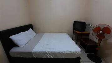 A small, dimly lit room with a single bed, a television, and a standing fan.