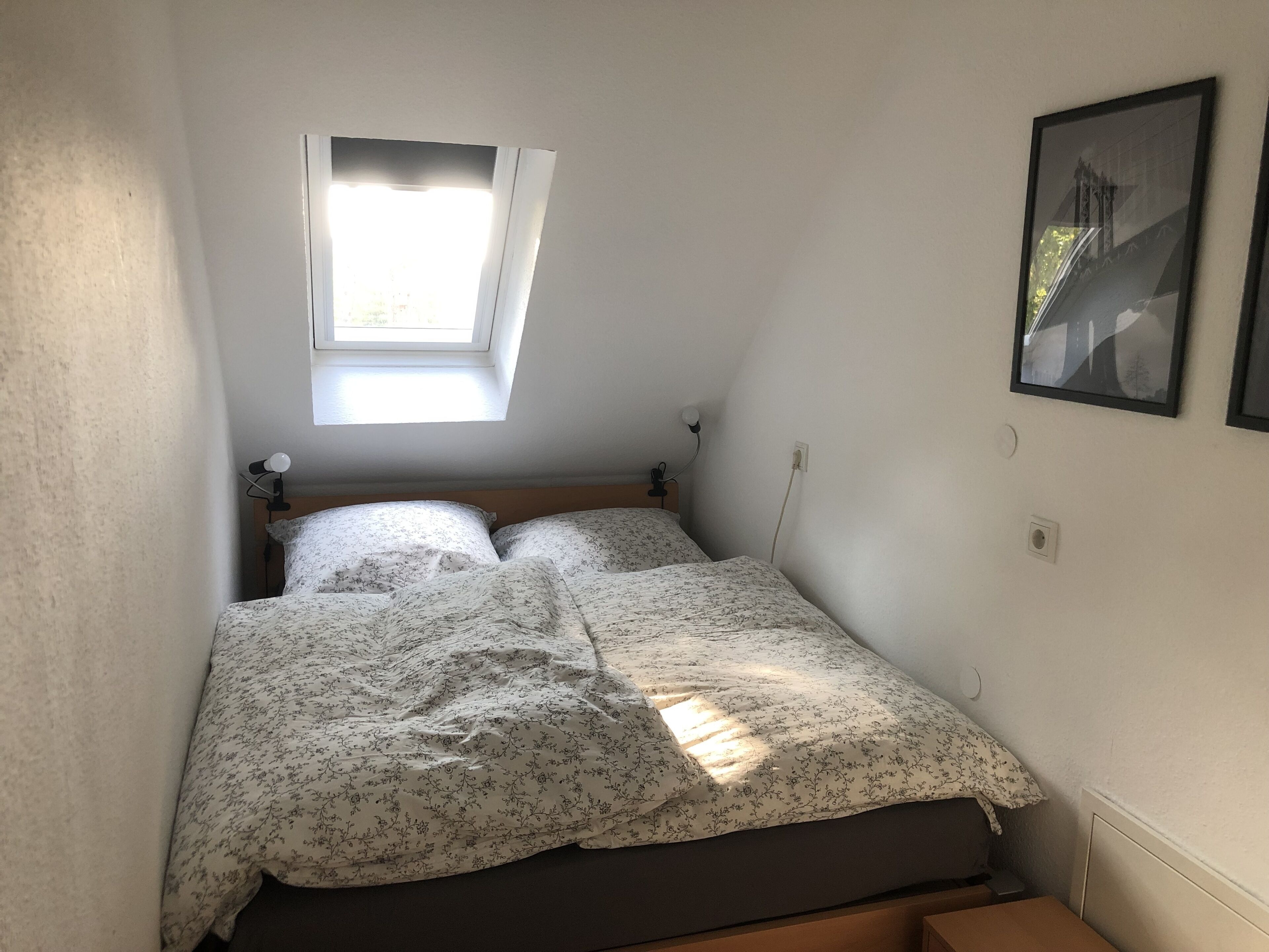 A small, single-bed room with a slanted ceiling, a skylight, a bedside lamp, and a framed picture on the wall.