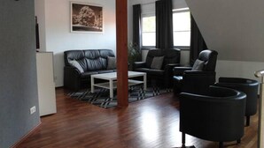 Una sala de estar moderna con un sofá de cuero negro, una mesa de centro blanca y sillones negros. Hay una ventana grande con cortinas, un reloj en la pared y un cuadro enmarcado sobre el sofá.
