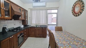 A compact kitchen with wooden cabinets, a gas stove, and a sink. There is a small dining area with a patterned tablecloth and a chair. - COMFORTABLE THREE BEDROOM APARTMENT WITH WIFI & LOCATED IN THE CENTRE  (QAWRA)