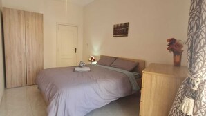 A bedroom with a bed, a wooden wardrobe, a bedside table with a lamp, and a small painting on the wall. - COMFORTABLE THREE BEDROOM APARTMENT WITH WIFI & LOCATED IN THE CENTRE  (QAWRA)