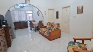 A living room with a sofa, a wooden cabinet, a dining area with a table and chairs, and a kitchen visible through an archway. - COMFORTABLE THREE BEDROOM APARTMENT WITH WIFI & LOCATED IN THE CENTRE  (QAWRA)
