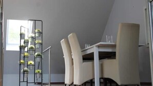 A dining area with a white table and four chairs, a black plant stand with multiple potted plants, and a window providing natural light. - Schöne Eigentumswohnung mit Terrasse für 2 Pers (Bippen)