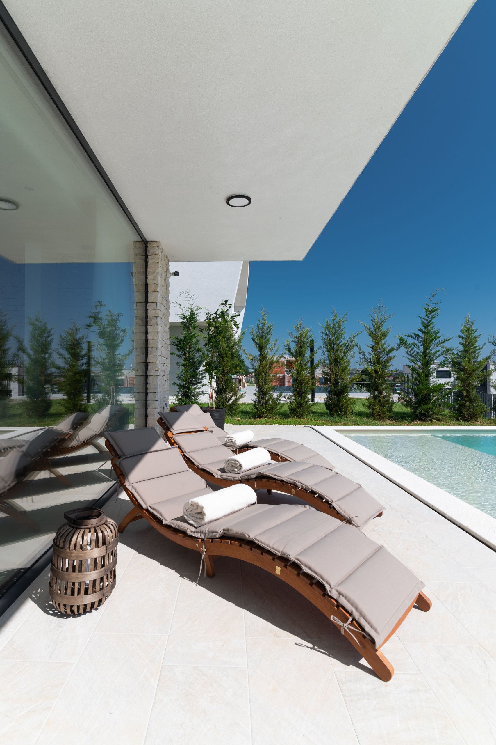 A modern outdoor lounge area with a pool, featuring a curved wooden lounge chair with cushions, a decorative lantern, and a clear view of the surrounding landscape.