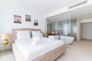 A modern hotel room with a large bed, a glass shower enclosure, and a view of the sea.