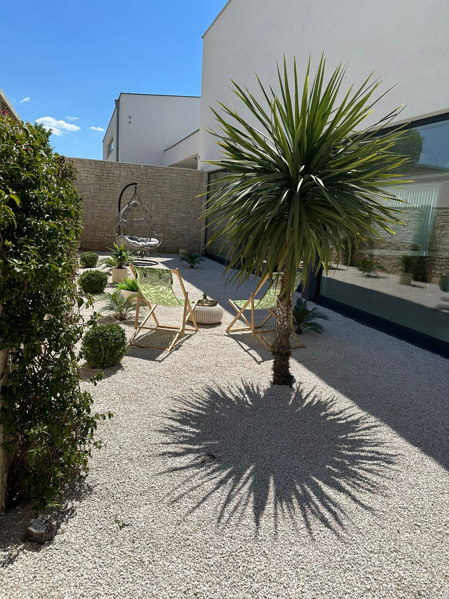 Un patio moderno al aire libre con una mecedora colgante, una pequeña palmera y suelo de grava.