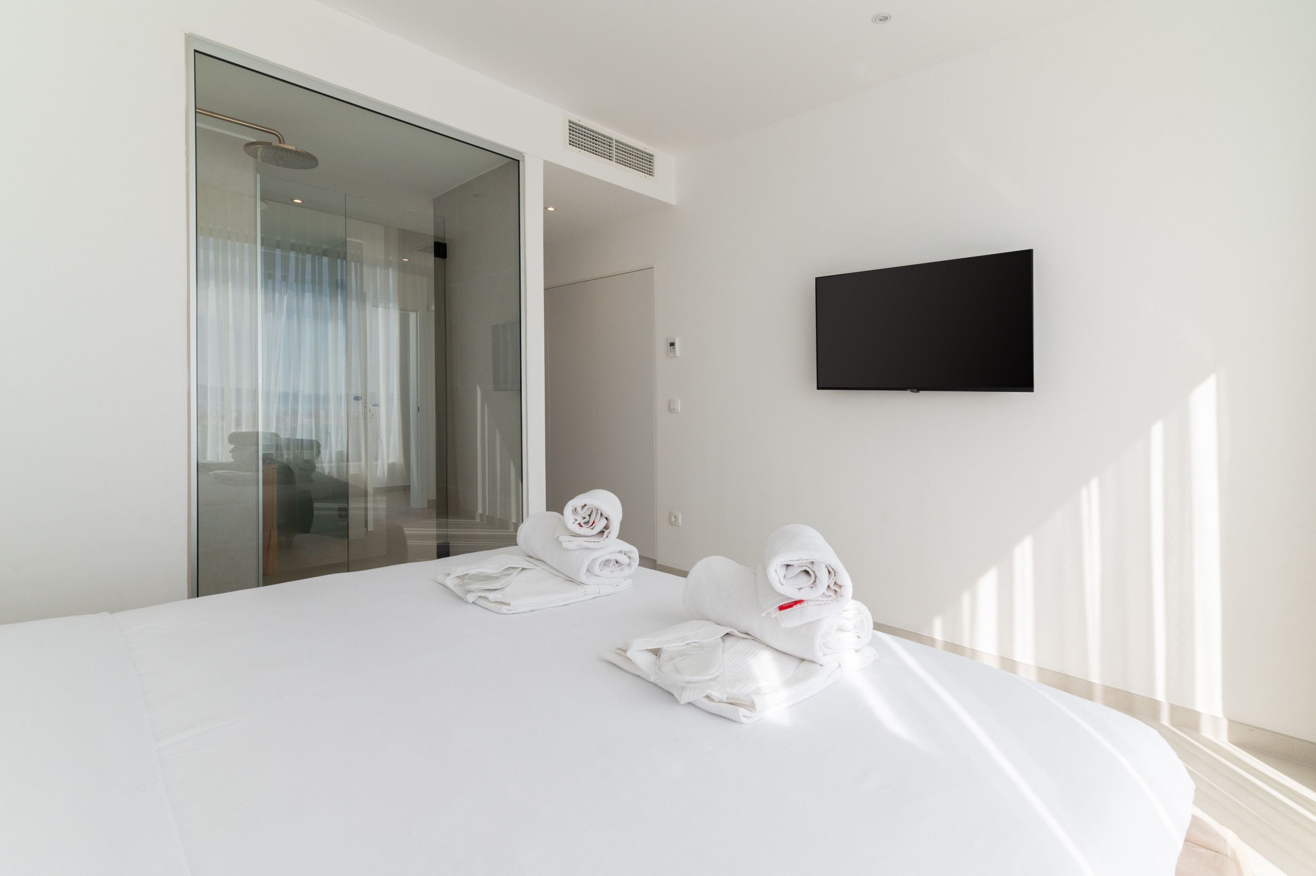 A modern hotel room with a large bed, a flat-screen TV mounted on the wall, and a glass door leading to a bathroom.