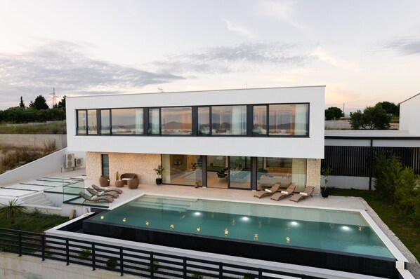 A modern two-story house with a swimming pool, outdoor seating, and a view of the surrounding landscape.