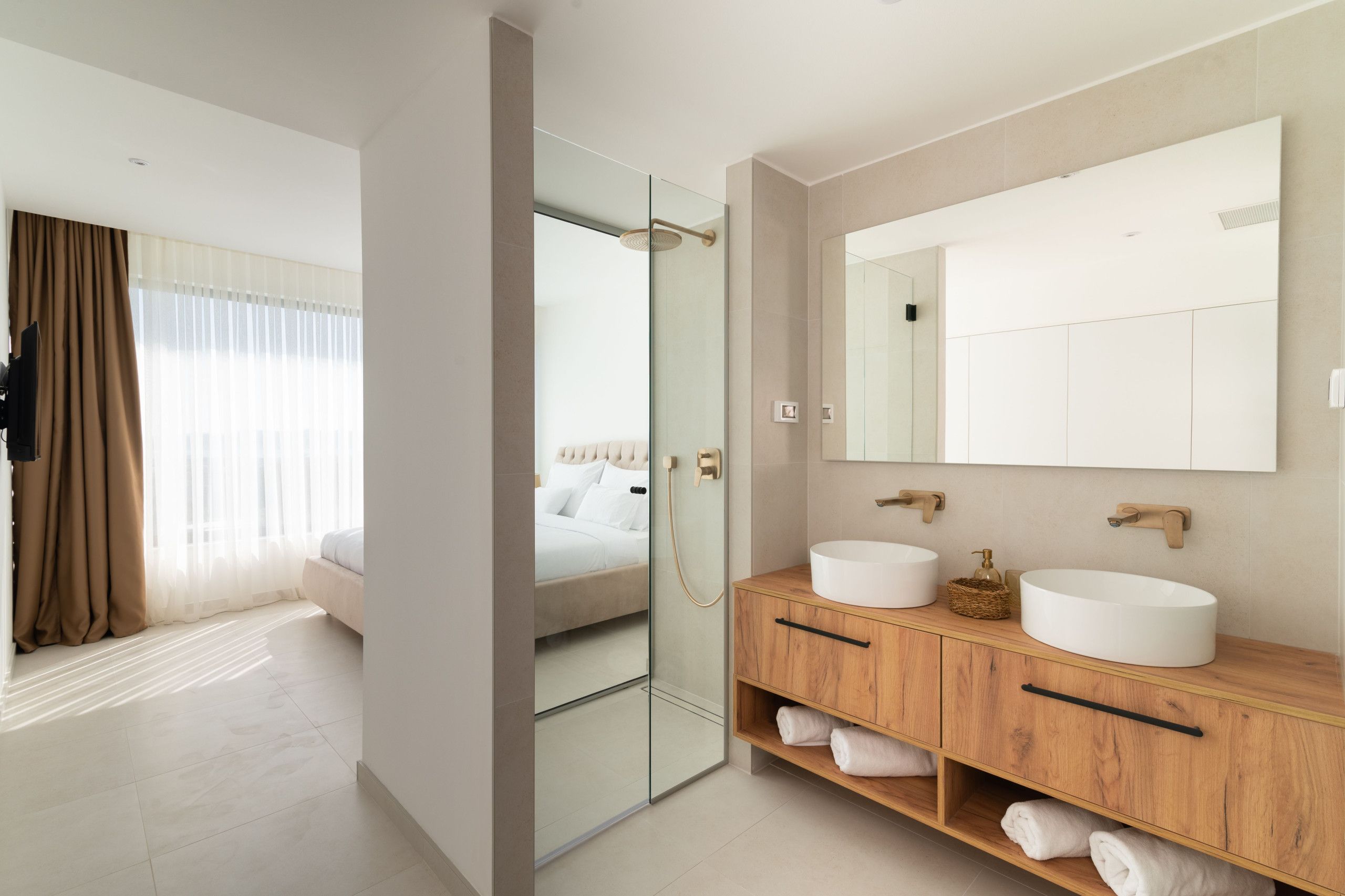 A modern hotel room with a large bed, a glass shower enclosure, a wooden vanity with two sinks, and a large mirror.