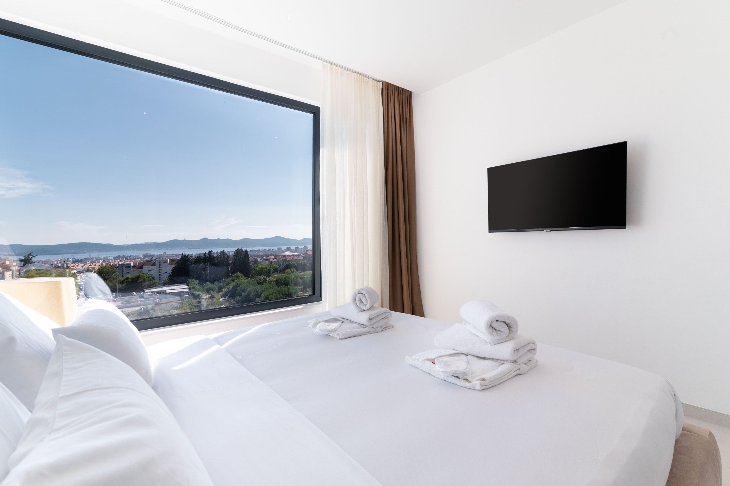 A hotel room with a large window offering a city view, a flat-screen TV mounted on the wall, and neatly folded towels on the bed.