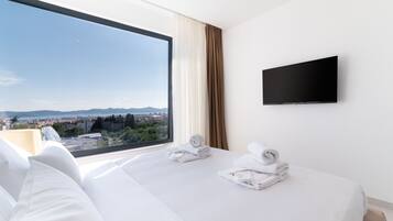 A hotel room with a large window offering a city view, a flat-screen TV mounted on the wall, and neatly folded towels on the bed.