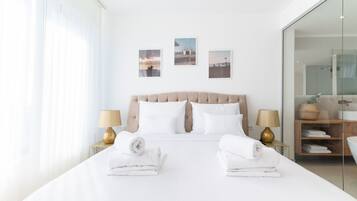 Una habitación de hotel moderna con una cama grande, dos mesitas de noche con lámparas, un box de ducha de vidrio y tres cuadros en la pared.