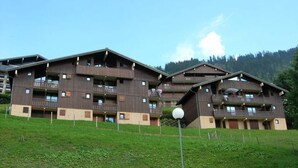 A multi-story wooden building with balconies, set against a backdrop of green hills and a clear sky.