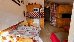 A compact kitchen with wooden cabinets, a microwave, and a refrigerator. There is a dining table with a checkered tablecloth and wooden chairs.