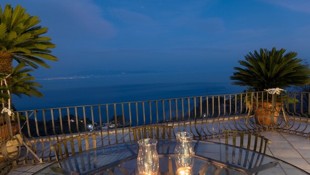Une table en verre décorée de bougies, des chaises en métal et une vue sur l’océan au crépuscule.