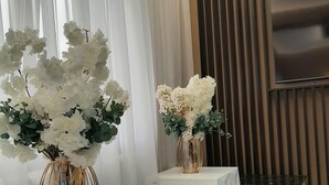 A modern room with two white tables, each with a vase of white flowers. There is a flat-screen TV mounted on a wooden panel wall, and the room has a dark marble floor. - Pensiunea Ramina (Adjud)