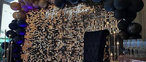 Una habitación con un arco de globos negros y dorados, un muro espejado y una mesa con copas.