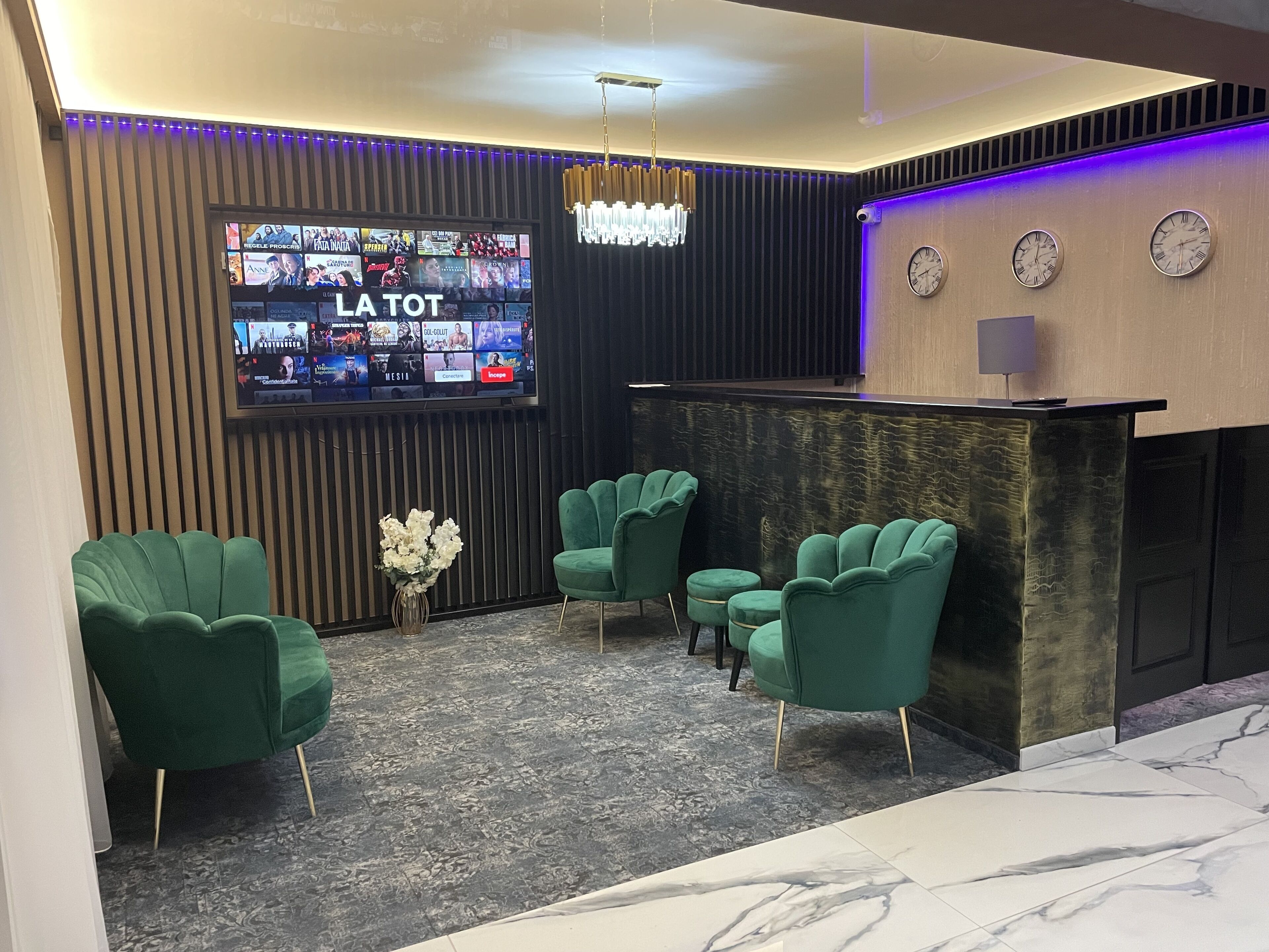 A modern reception area with a flat-screen TV displaying various channels, a chandelier, and green upholstered chairs.