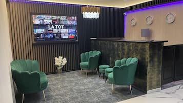 A modern reception area with a flat-screen TV displaying various channels, a chandelier, and green upholstered chairs.