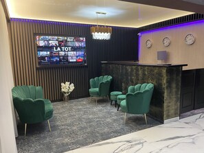 A modern reception area with a flat-screen TV displaying various channels, a chandelier, and green upholstered chairs.