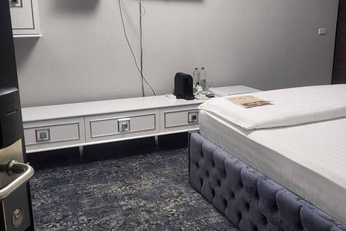 A hotel room with a bed, a flat-screen TV mounted on the wall, a small bedside table with a lamp, and a built-in wardrobe.