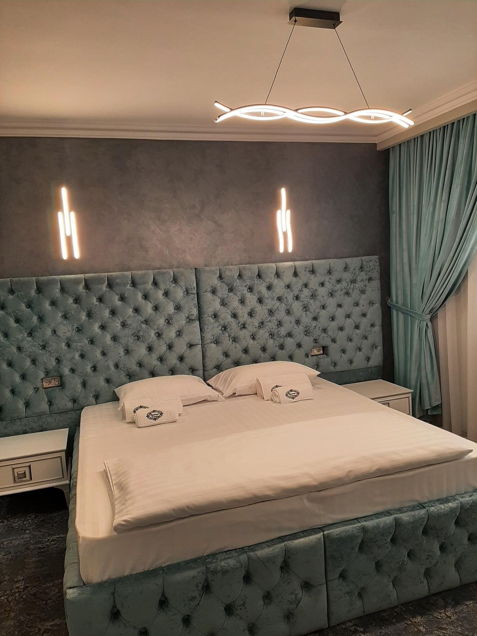 A bedroom with a tufted headboard, a bed with neatly folded towels, and a modern light fixture.