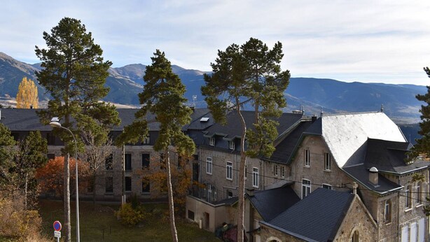 A residential area with stone buildings, large trees, and a mountainous backdrop.