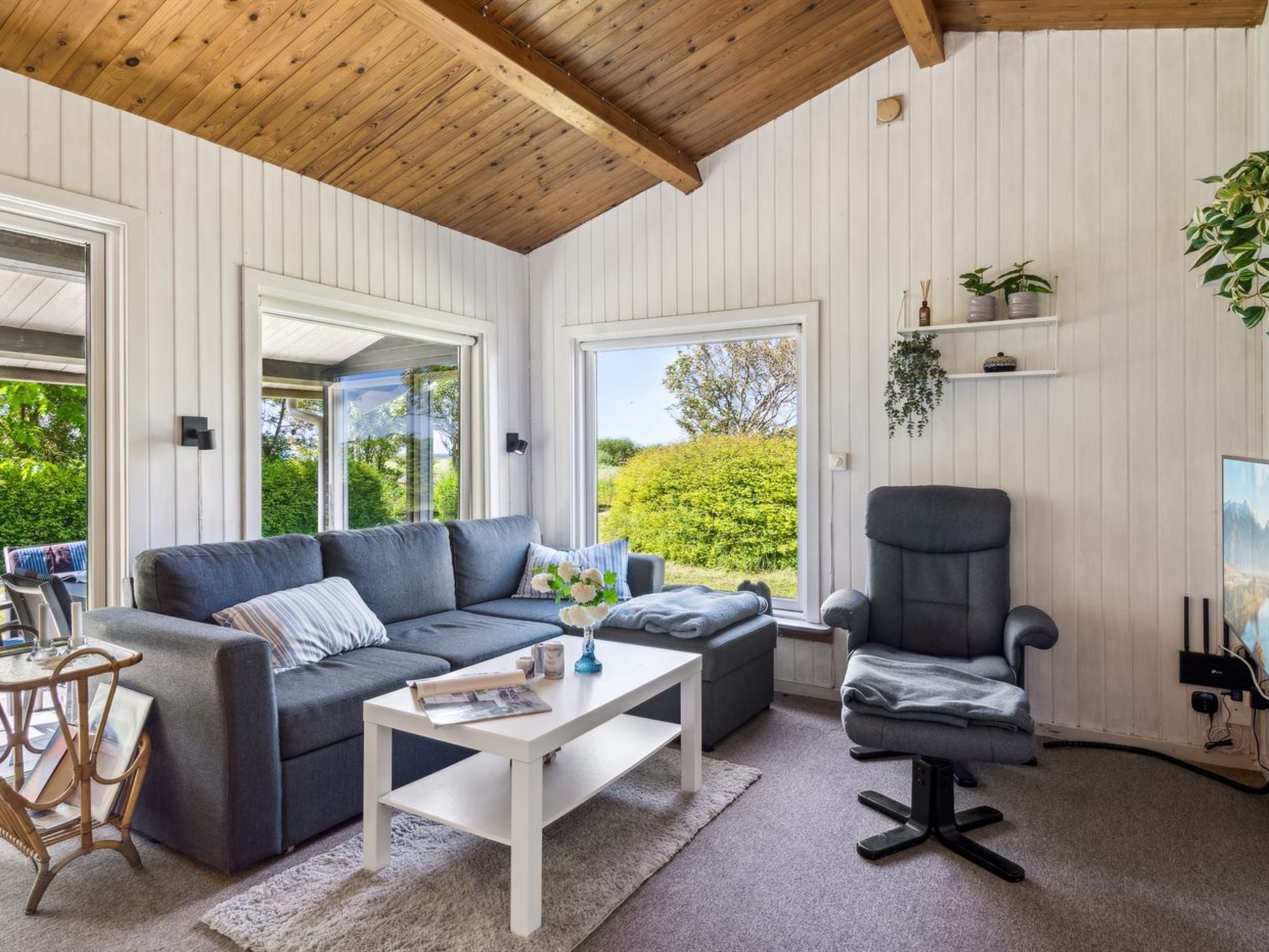Una sala de estar moderna con un sofá gris, una mesa de centro blanca y un sillón gris. Amplias ventanas ofrecen vistas a zonas verdes al exterior.