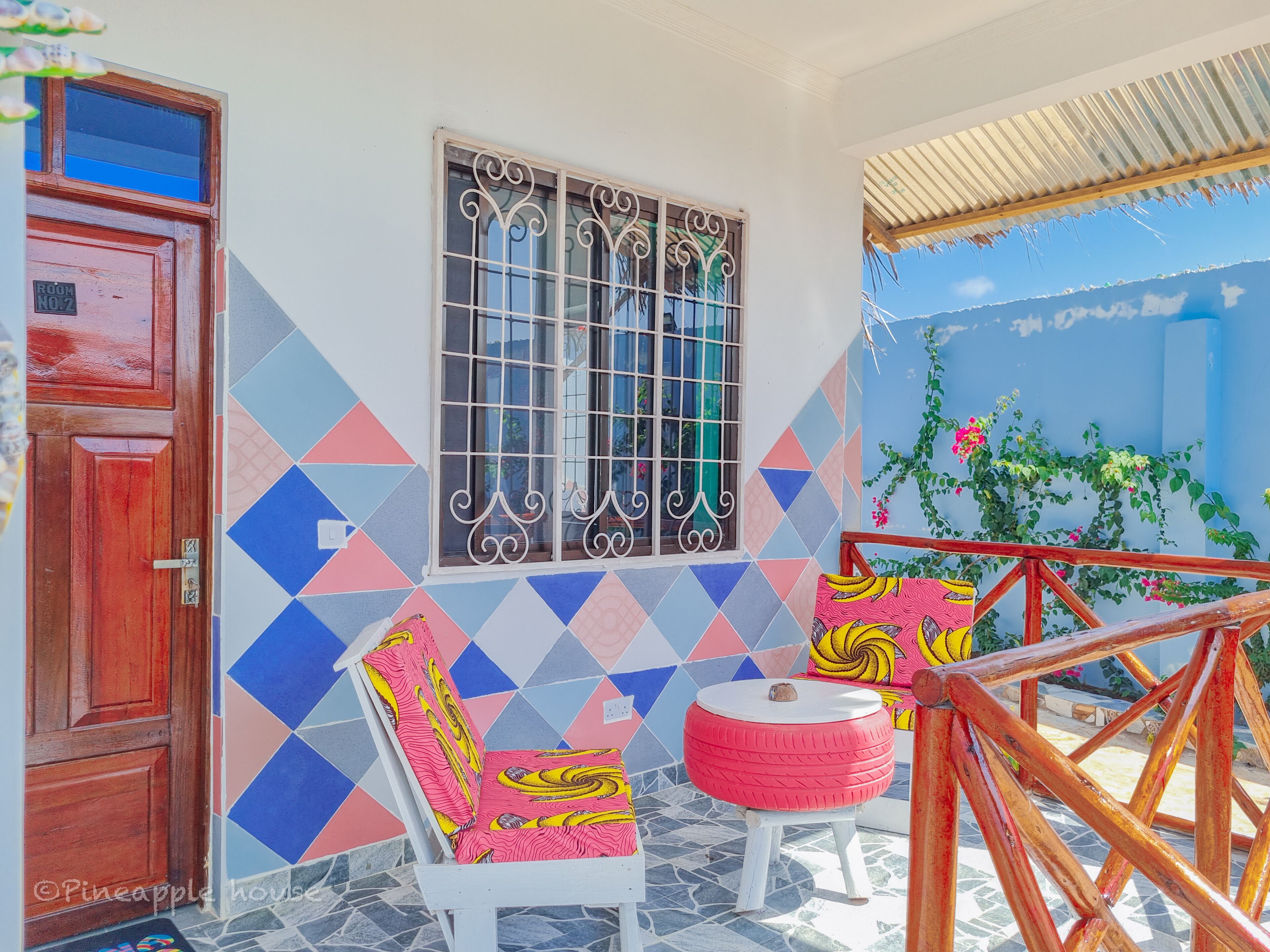 A room with a tiled floor, a wooden door, a window with decorative bars, a chair with a colorful cushion, a small stool, and a wooden chair frame.