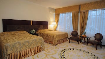 A hotel room with two beds, a sitting area with chairs, a small table with fruit, and a large window with curtains.