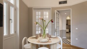 A dining area with a round table set for four, surrounded by white chairs. There are floral arrangements on the table, and a kitchen is visible in the background. - Acropolis Maisonette 22 (Athina)