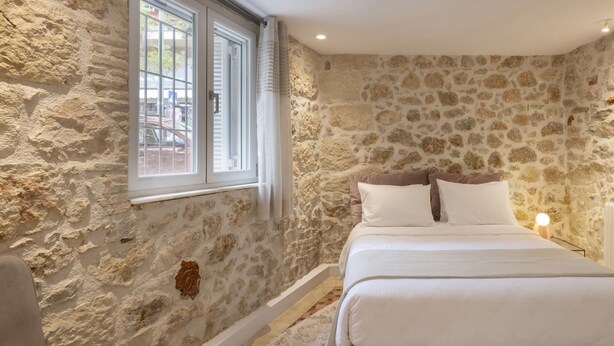 A bedroom with a stone wall, a bed with white bedding, a window with blinds, and a nightstand with a lamp. - Acropolis Maisonette 22 (Athina)