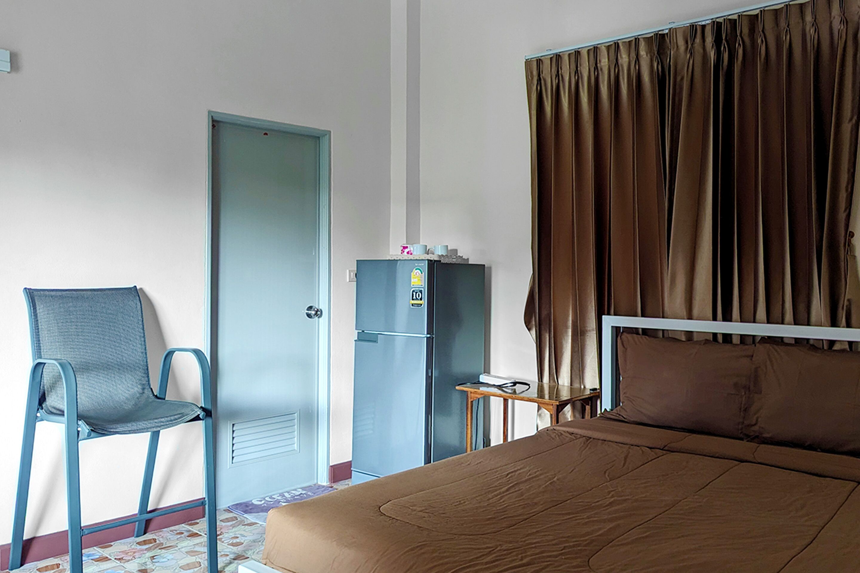 Una habitación de hotel con cama, una silla, una mesita, un refrigerador y una puerta.