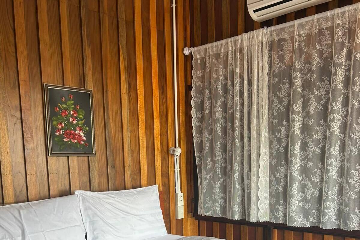 A neatly made bed with white linens, a floral painting on the wall, and a window with a patterned curtain.