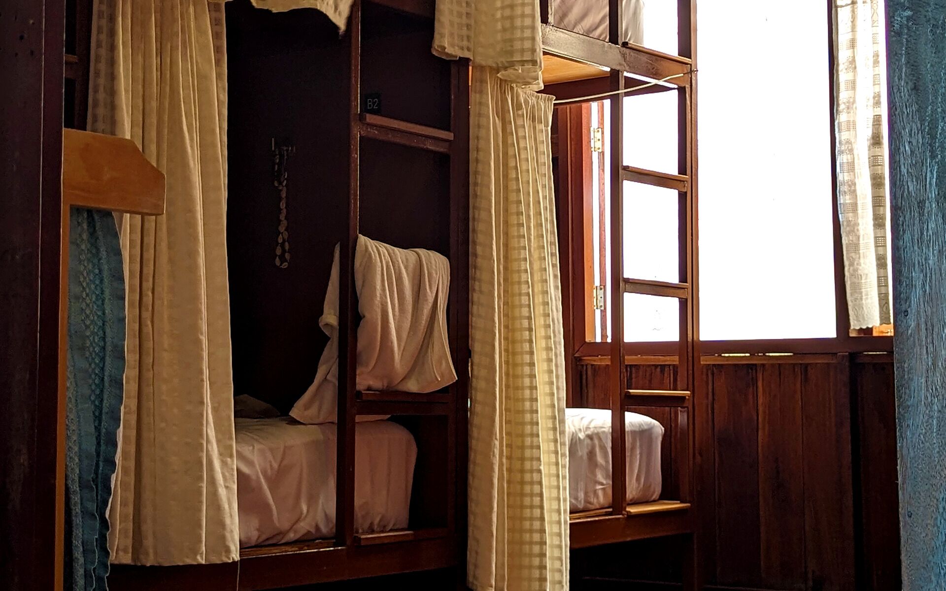 A room with two wooden bunk beds, a window with curtains, and a wall-mounted chain.