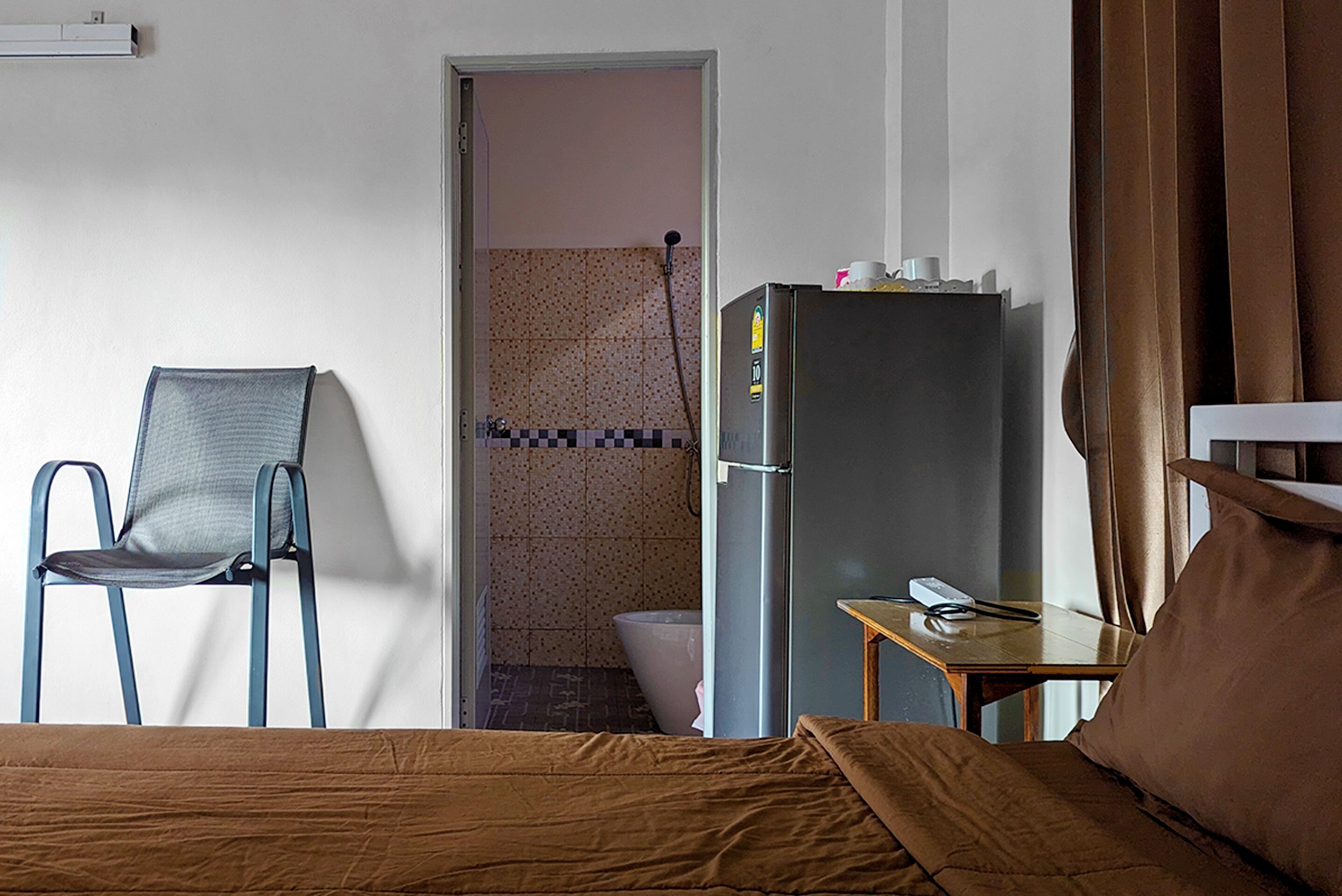 A hotel room with a bed, a chair, a refrigerator, a small table, and a bathroom visible through an open door.