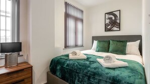 Et pænt hotelværelse med en seng, et natbord med lampe, et vindue med persienner og et billede på væggen.