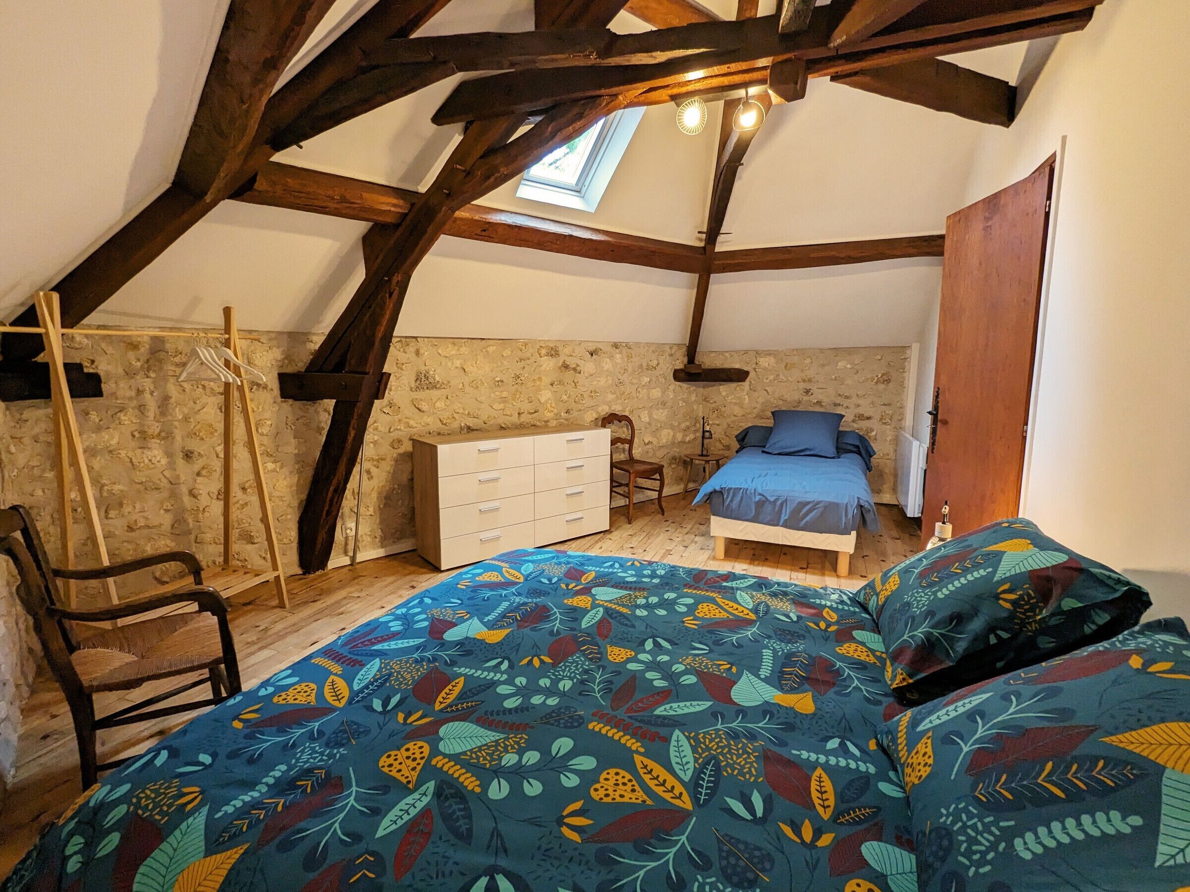 Ein Schlafzimmer mit schrägverdeckter Decke, einem Bett mit farbigem Blättermuster-Überwurf, einem hölzernen Schminktisch, einem Stuhl und einem kleinen Tisch.
