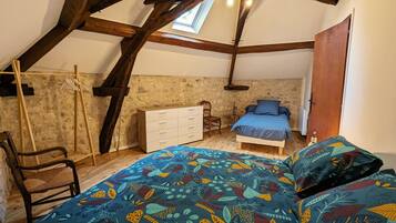 Ein Schlafzimmer mit schrägverdeckter Decke, einem Bett mit farbigem Blättermuster-Überwurf, einem hölzernen Schminktisch, einem Stuhl und einem kleinen Tisch.