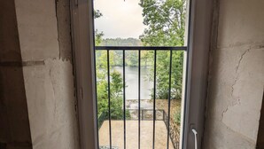 Uitzicht door een deur met een metalen hek, op een balkon met uitzicht op een wateroppervlak met bomen en een verre oever.