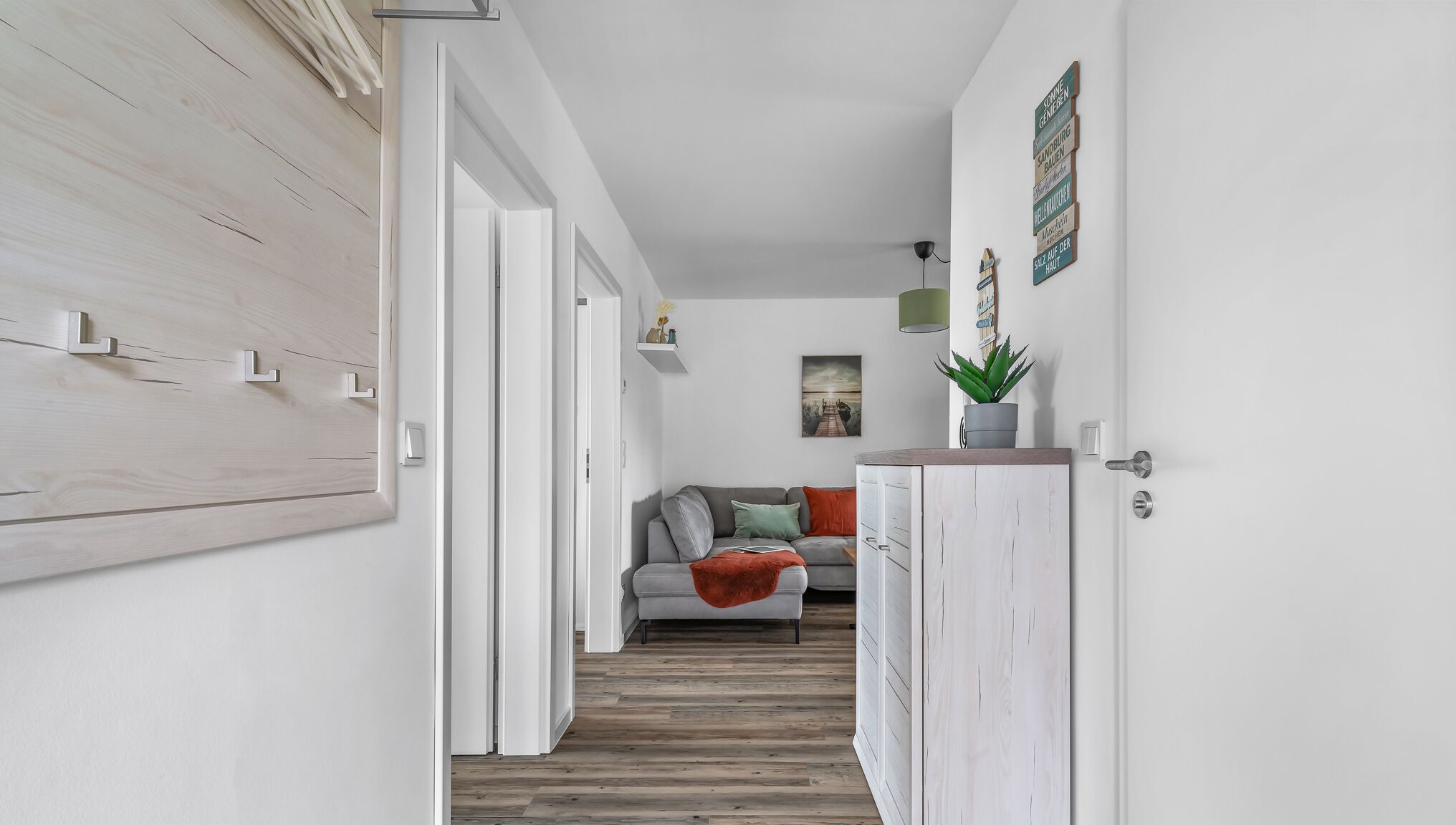 Ein Flur mit Holzboden, einem grauen Sofa mit Kissen, einer roten Decke und einem weißen Schrank mit Pflanze.