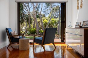 A room with a wooden floor, two chairs, a small round table, and a cabinet. There is a large window with a view of trees and a garden. - Golden Sands Rye - 1 bedroom for 2 guests - Tranquil, spacious retreat for two. Close to Tyrone beach. (Rye)